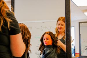 A young woman is working on the hair of a mannequin while standing in front of a mirror.